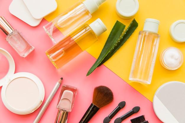 Various skincare and makeup products arranged on a colorful background, including bottles of serum, aloe vera, compact powders, and makeup brushes.