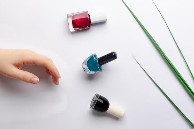 A child's hand reaching towards various nail polish bottles arranged artistically with thin green reeds on a white background.